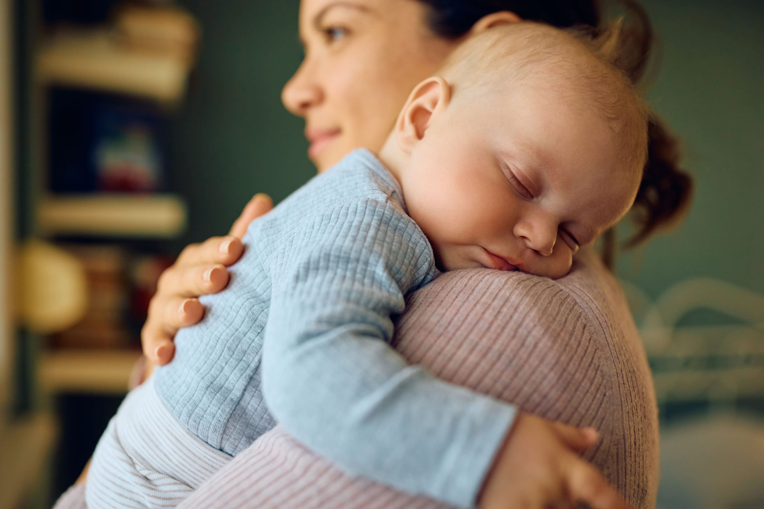 A mother holds her sleeping baby gently on her shoulder. The baby, dressed in a light blue ribbed outfit, rests peacefully with eyes closed and a calm expression. The mother, partially visible and wearing a pink top, looks content and focused, creating a warm and nurturing scene.